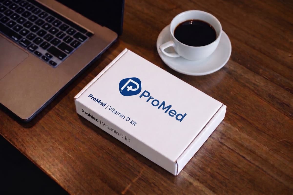 A ProMed Vitamin D home collection kit on a desk beside a laptop and a cup of coffee